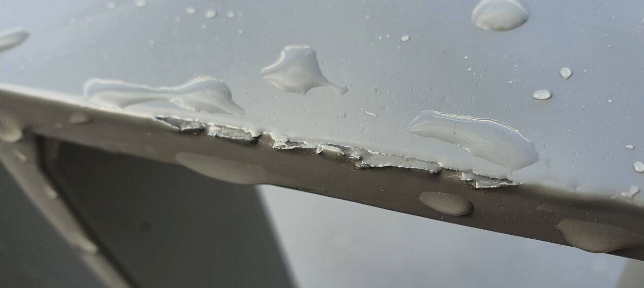 Grau lackierte Metalloberfläche mit abgeplatzter Kante und Wassertropfen.
