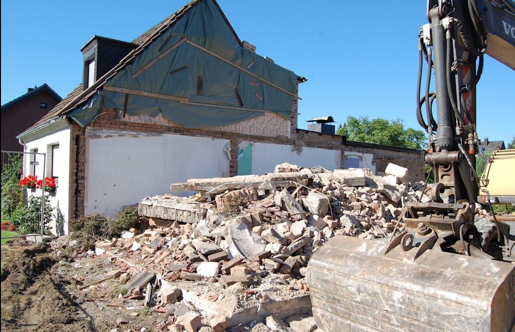 Zertrümmerte Hausruine mit Bruchsteinmauerwerk, Bagger auf Baustelle vor blauem Himmel