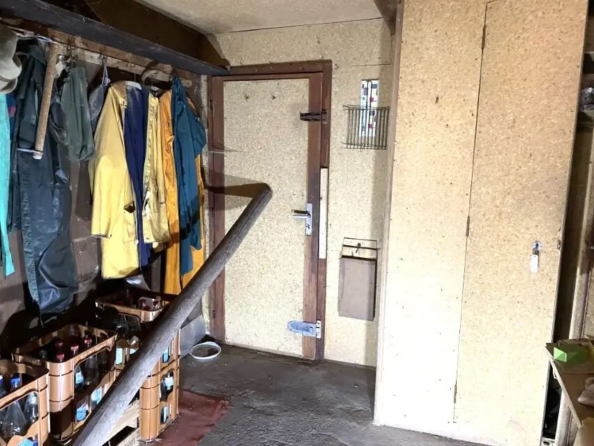Kellerflur mit Garderobe links, bunten Jacken, Getränkekisten unten, Tür in der Mitte.