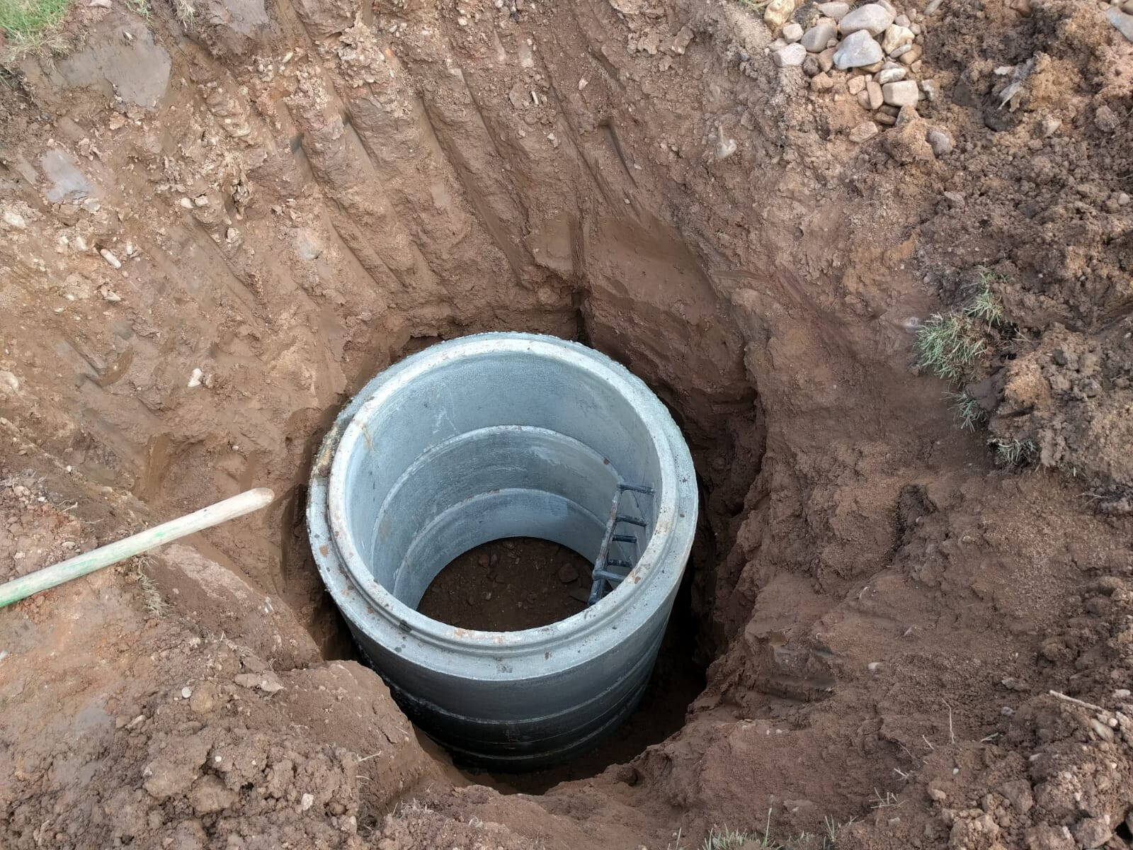 Runder Beton-Manhole in einer Erdgrube; Innenraum mit Leiter sichtbar.