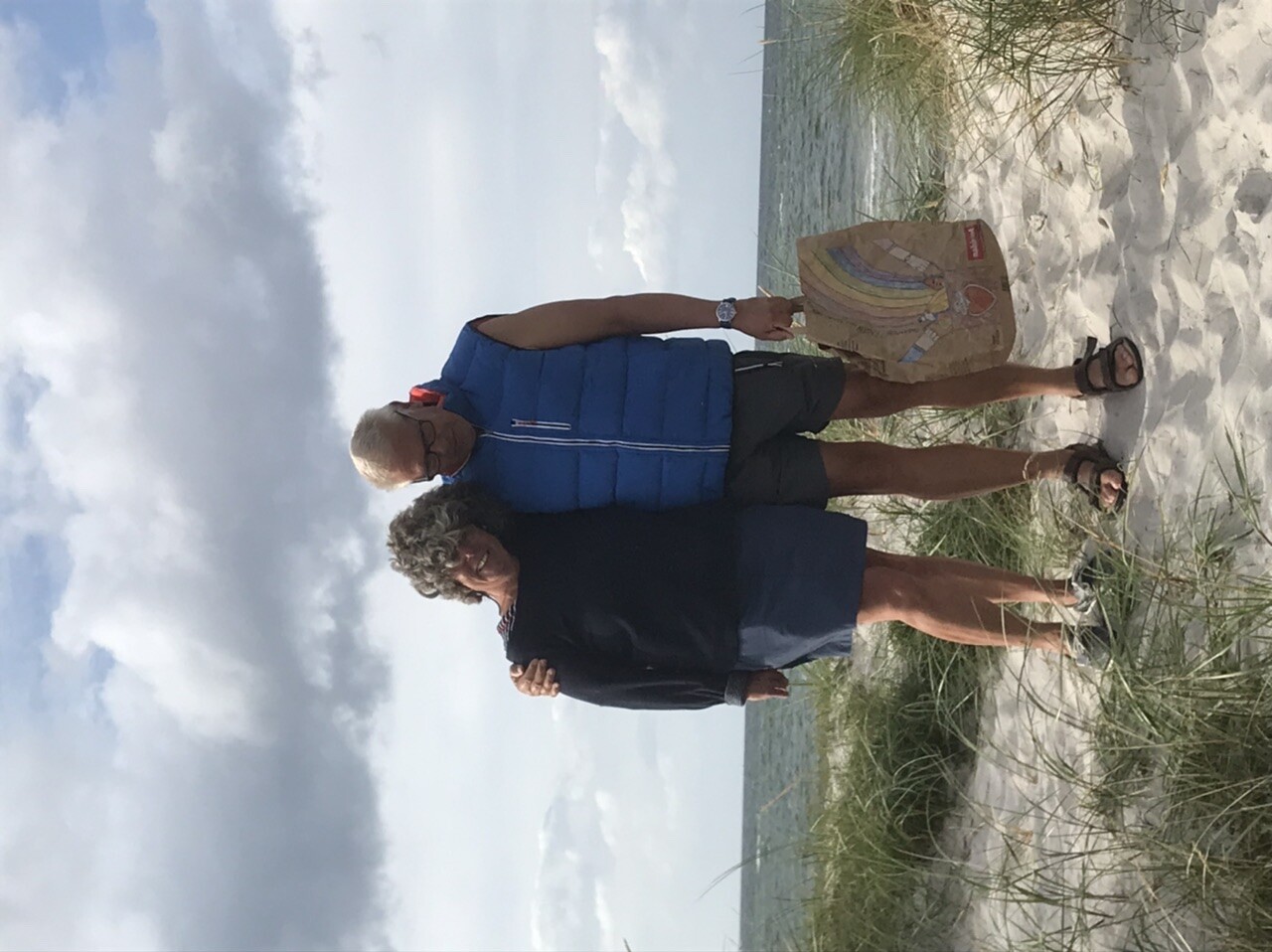 Paar am Strand auf Dünen, Meer im Hintergrund; bunte Tragetasche liegt da