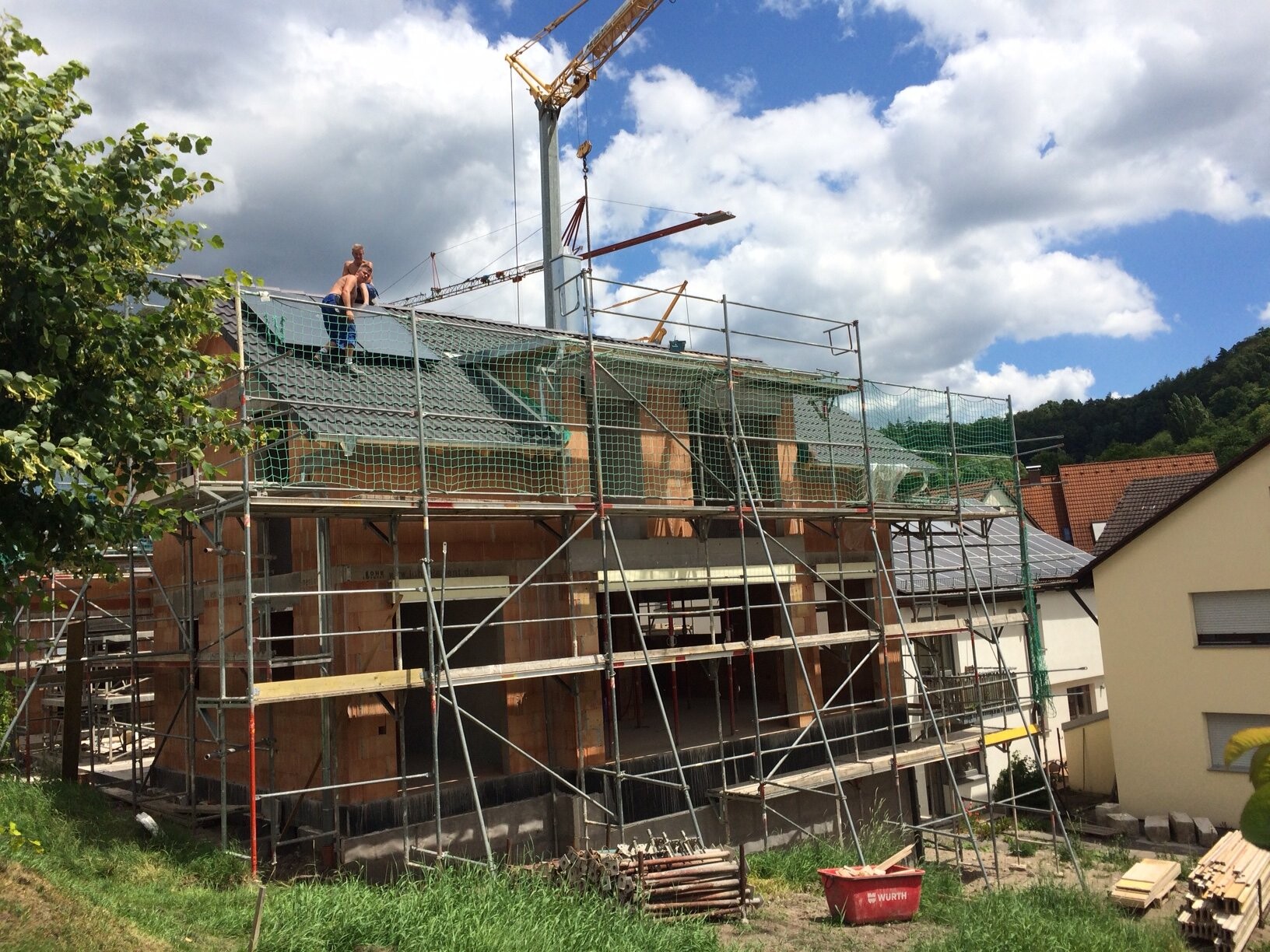 Baustelle mit Gerüst, zwei Arbeiter auf dem Dach, Kran im Hintergrund, umliegende Häuser
