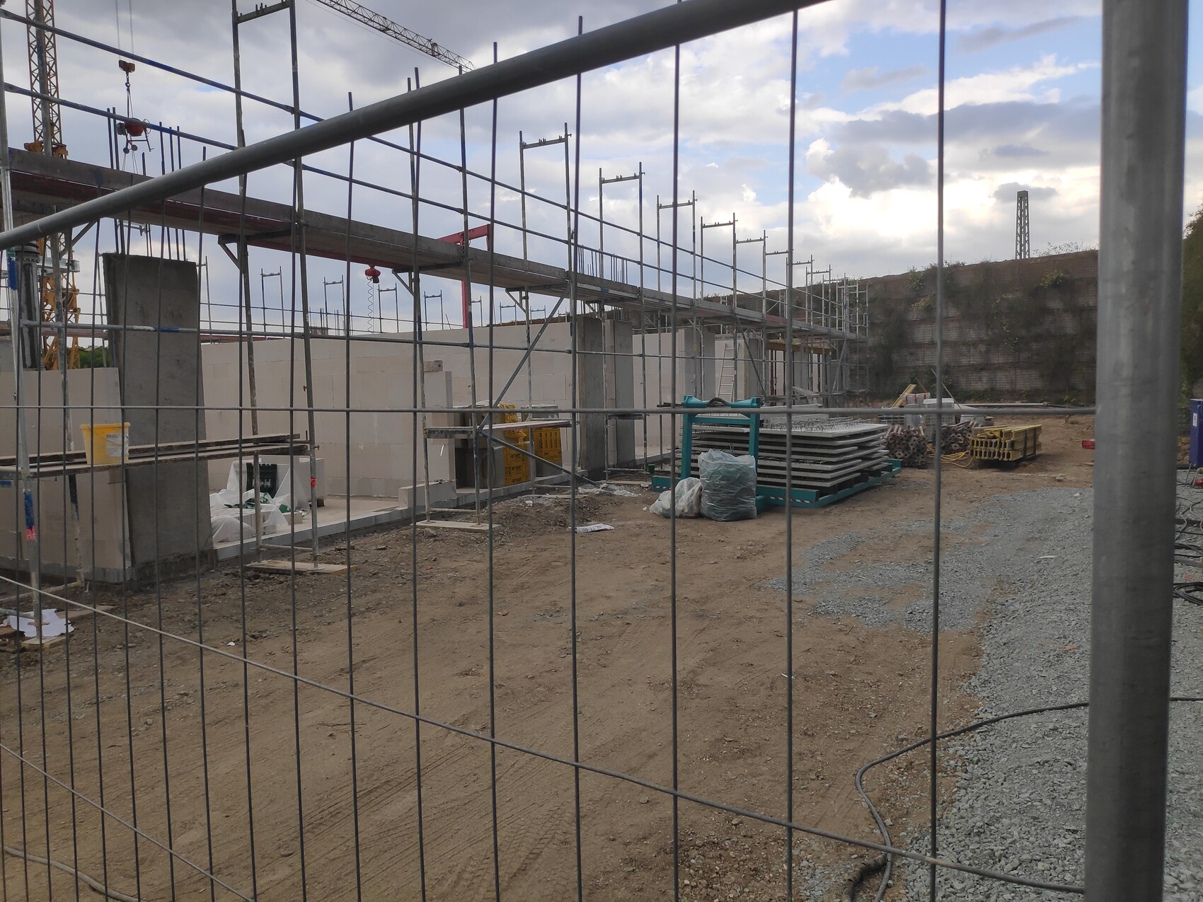 Baustelle mit Gerüst, Betonwänden und Baumaterialien hinter einem Metallzaun im Vordergrund.
