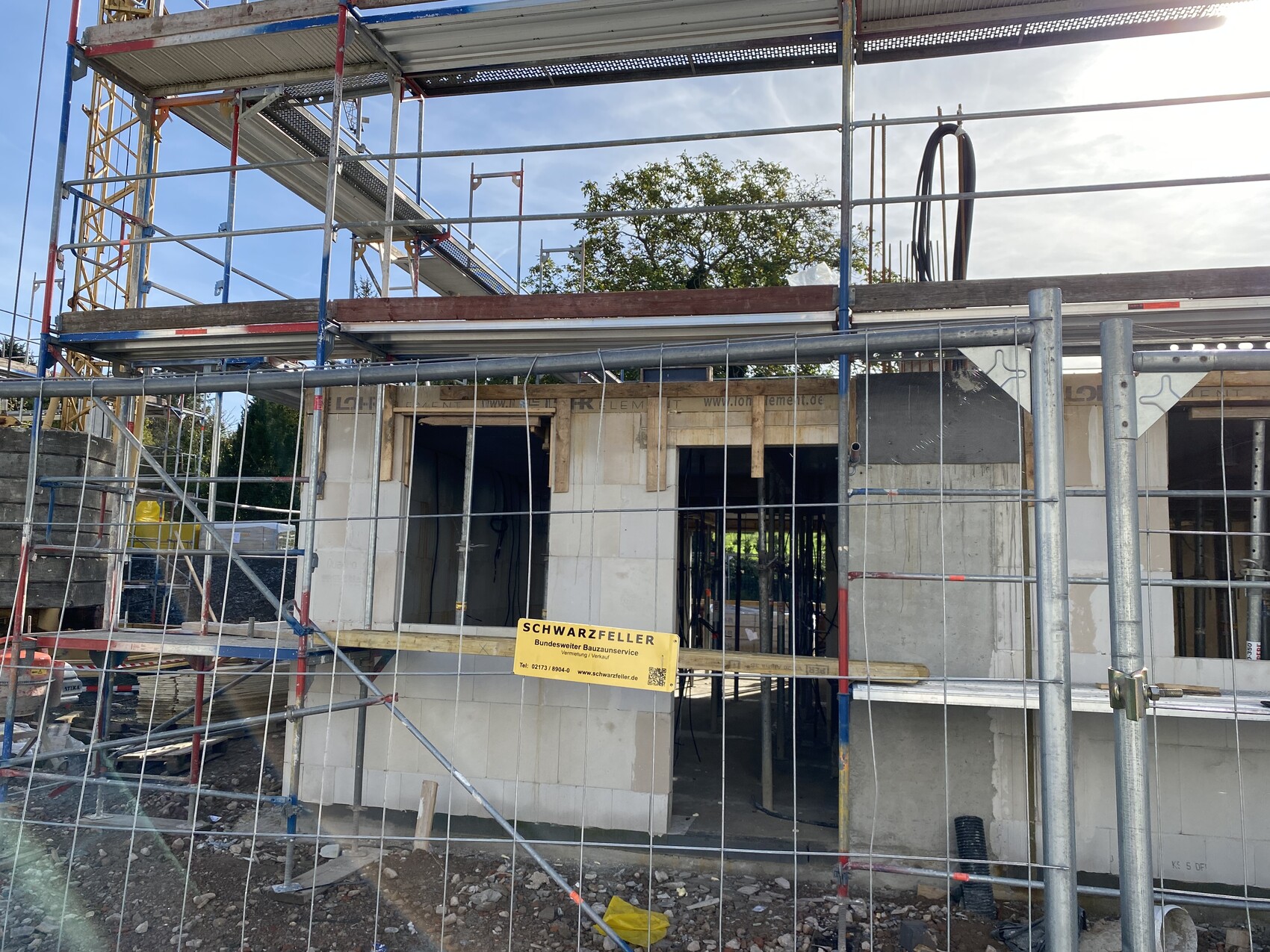 Baustelle mit Gerüst um Rohbau, Fenster- und Türöffnungen, Zaun im Vordergrund.