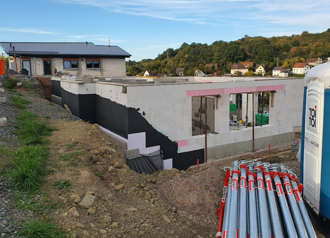 Halbfertige Baustelle mit weißem Mauerwerk, Stützen, Bauzubehör und Porta-Toilette.