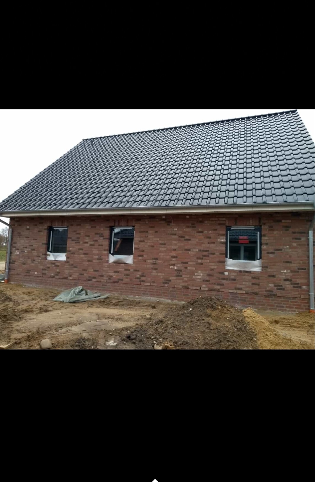 Backsteinhaus im Bau mit grauem Ziegeldach, drei Fenster abgedeckt, Baugrund davor.