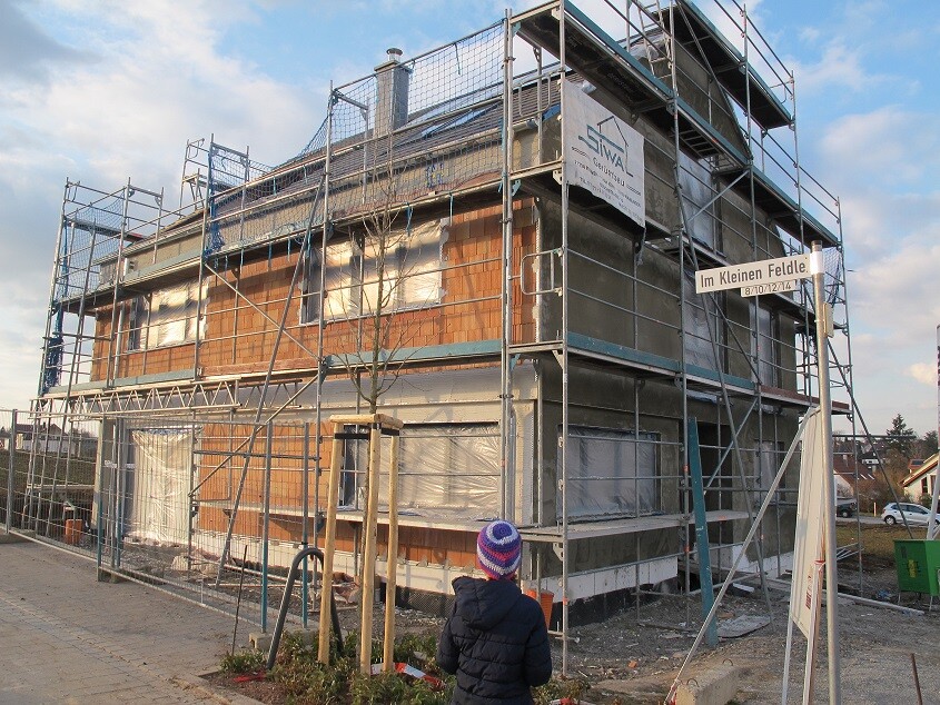 Mehrstöckiges Haus im Bau mit Gerüst; Kind in Winterjacke beobachtet die Baustelle.