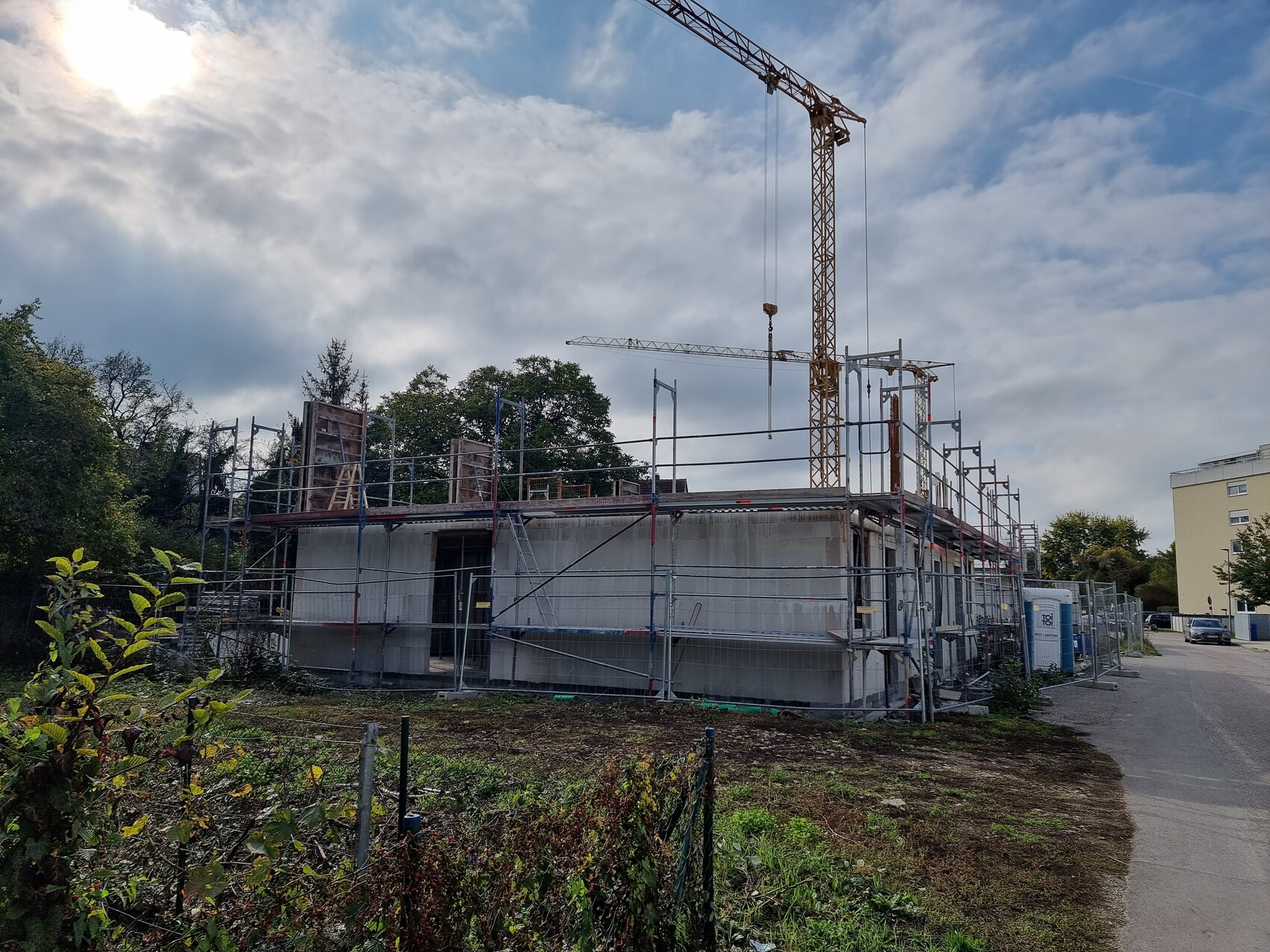 Baustelle mit Gerüst um unfertiges Gebäude; Kran ragt hoch, Bauzaun und Straßenszene.