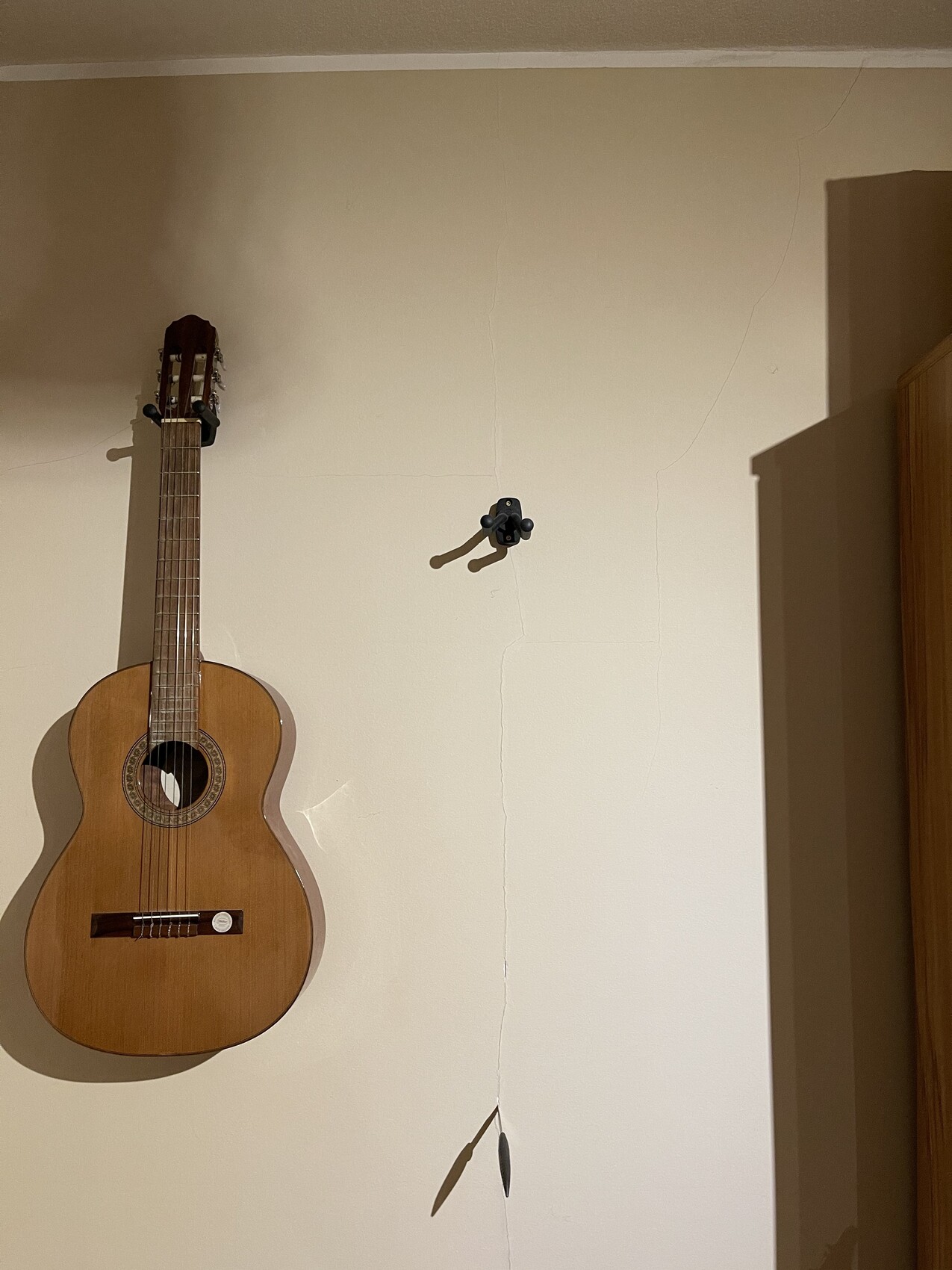 Akustikgitarre hängt an cremefarbener Wand mit Rissen; rechter Schrank sichtbar.