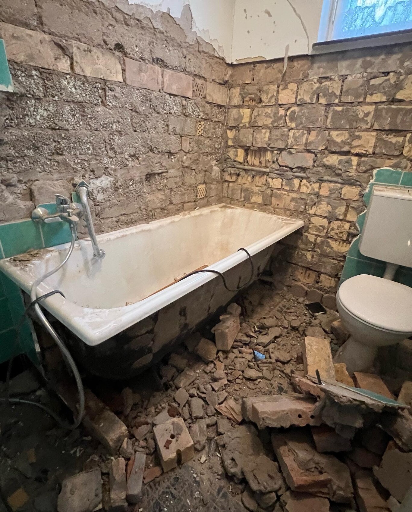 Verfallenes Badezimmer: freiliegende Steinmauern, Schutt auf dem Boden, Badewanne und Toilette.