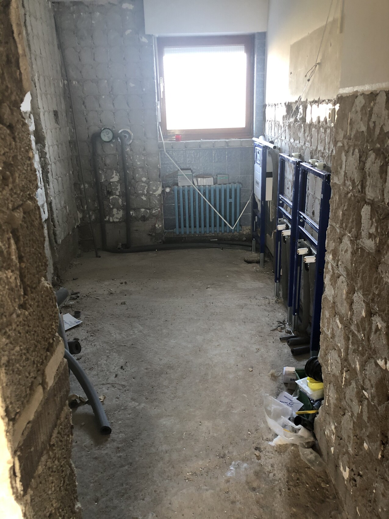 Kellerraum im Rohbau: Fenster, Heizkörper, Rohre und blaue Sanitärrahmen an der Wand.