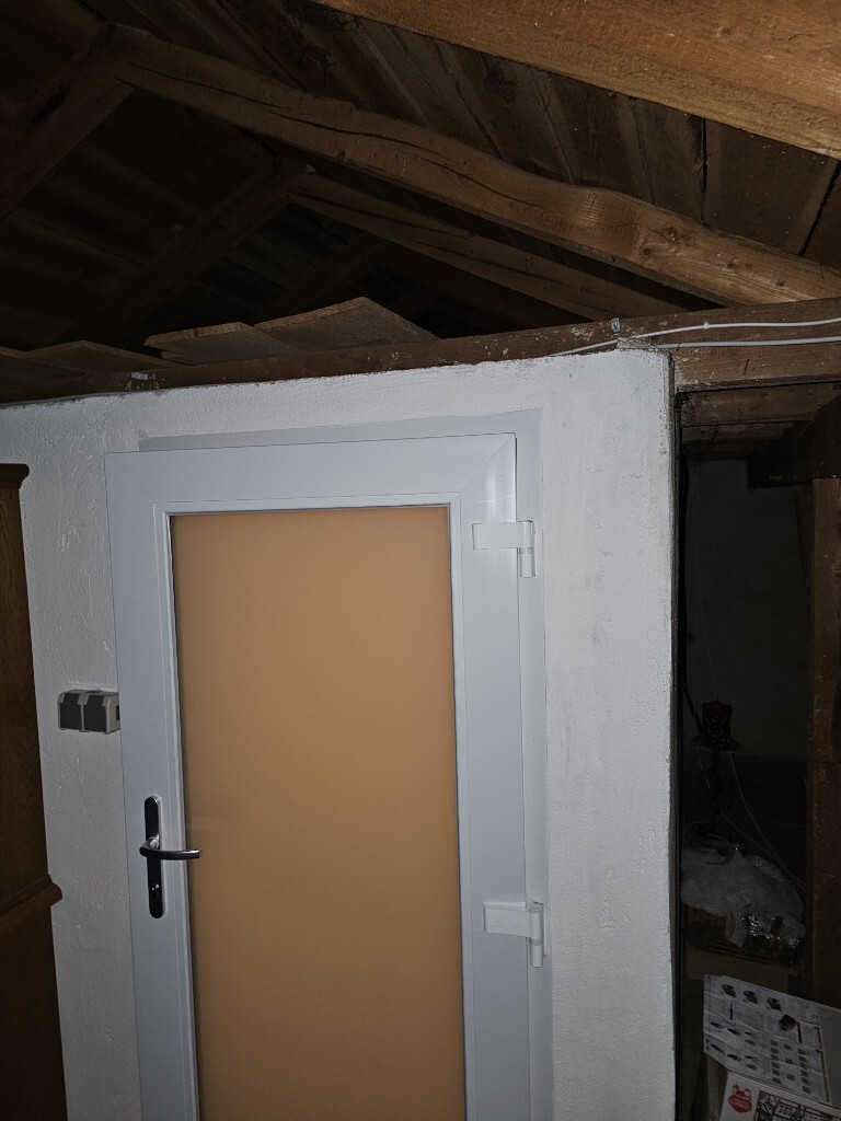 Weiße Tür mit orange getöntem Glas in einem Dachbodenraum; Holzbalken an der Decke.