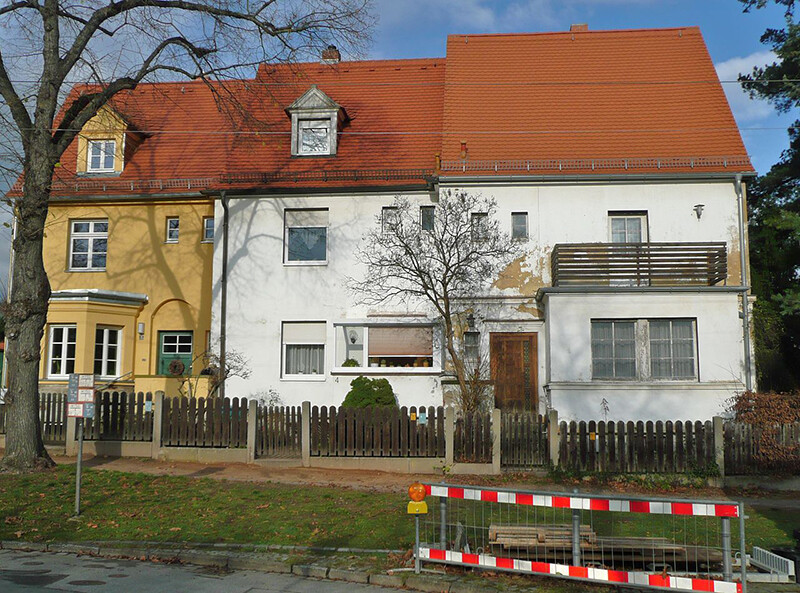 Straßenzug mit roten Ziegeldächern, gelbes Haus links, weißes Haus in der Mitte, Holzzaun, Bauzaun.