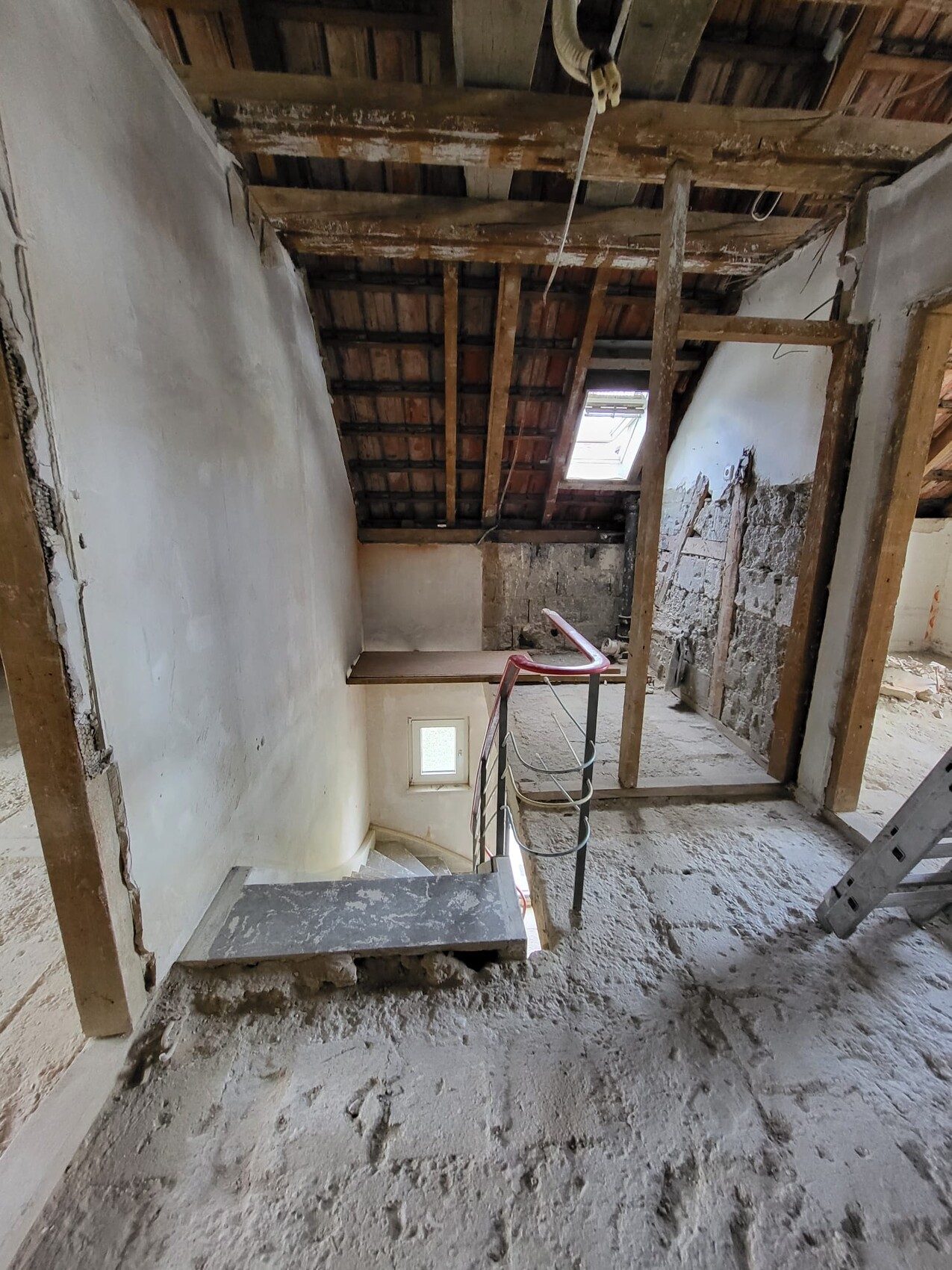 Dachgeschoss-Rohbau: Holzbalkendecke, rauer Boden, Stahlgeländer an Treppe, kleines Fenster.