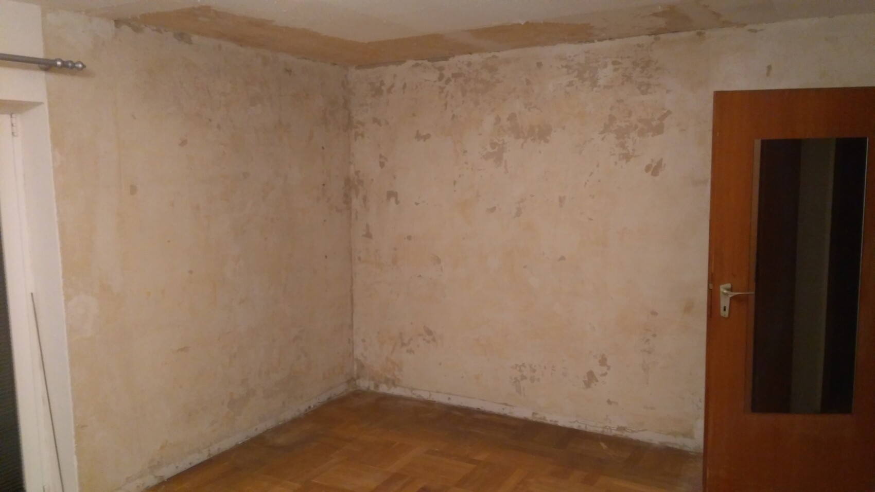 Verputzte Wände mit Abblätterungen, Ecke eines leeren Zimmers, Holzboden, Tür mit Glasfenster.