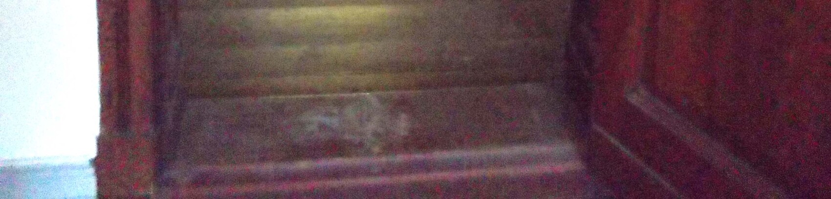 Dunkle Holztreppe von unten, rote Wand rechts, helle Wand links.