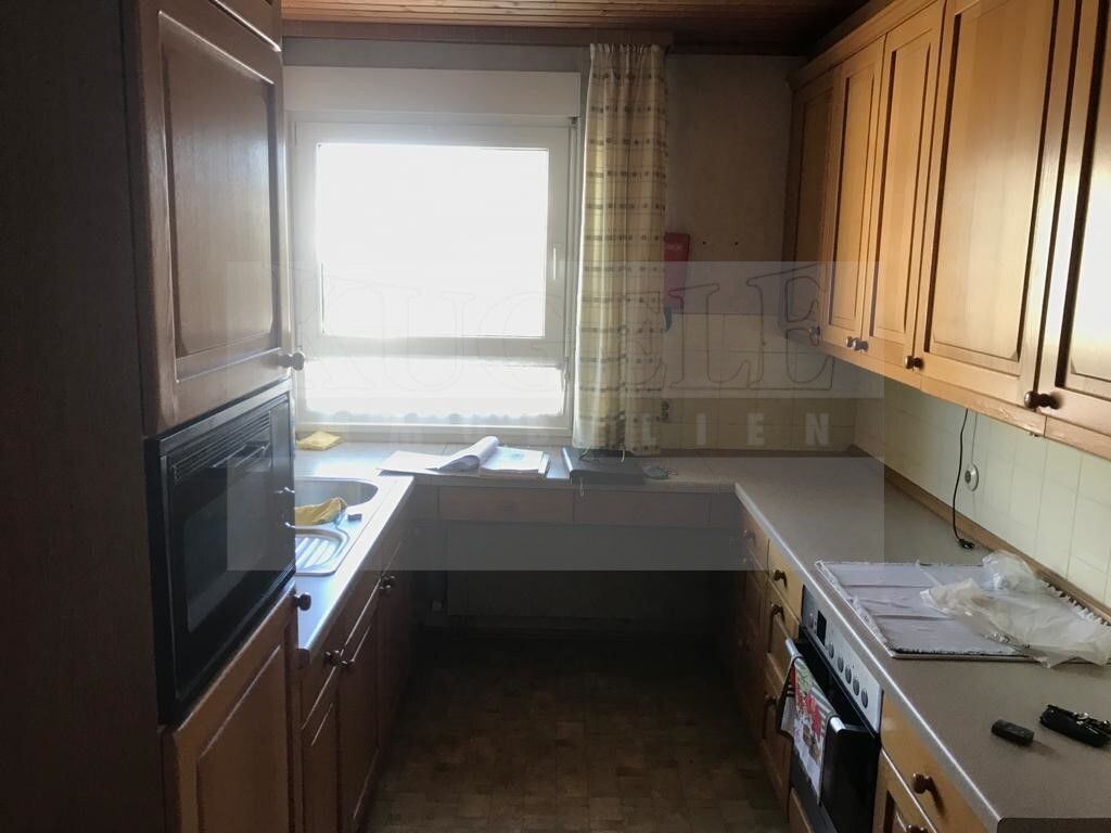 Kleine Küche mit Holzschränken, Spüle links, Ofen rechts, Fenster mit Vorhang im Hintergrund.