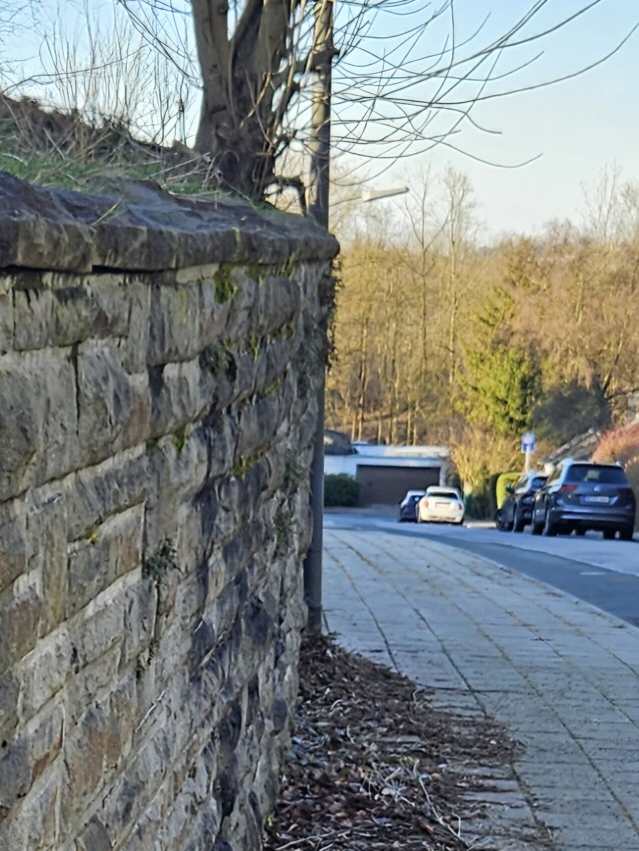 Blick entlang einer Steinmauer links, Gehweg rechts, geparkte Autos und kahle Bäume.