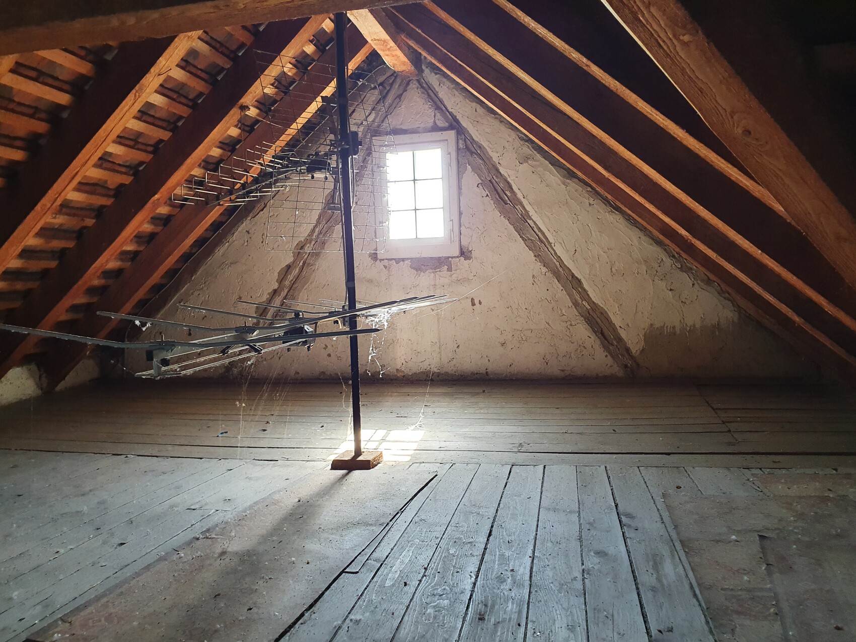 Dachboden mit Holzbalken, kleinem Fenster, Metall-Wäscheständer und staubigem Boden.