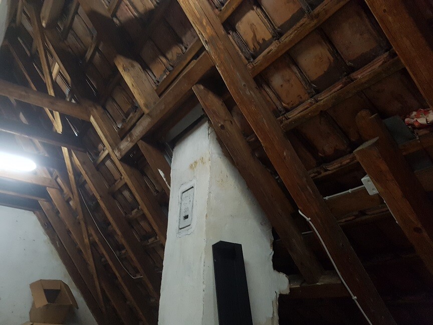 Dachboden mit sichtbaren Holzbalken, zentrale weiße Schornsteinwand und wenig Licht.