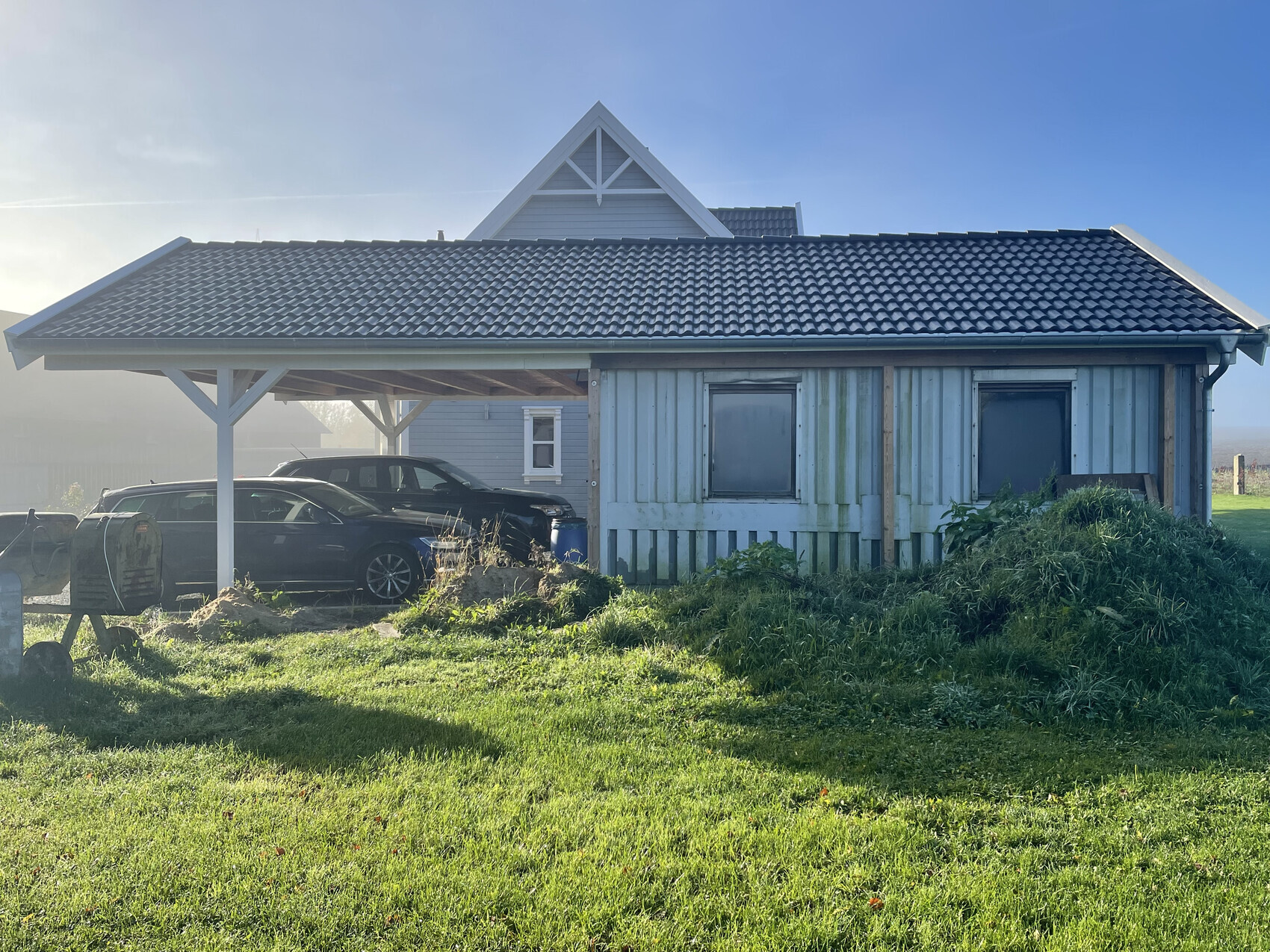 Kleines blaues Holzhaus mit Carport; zwei Autos davor und grüne Wiese im Vordergrund.