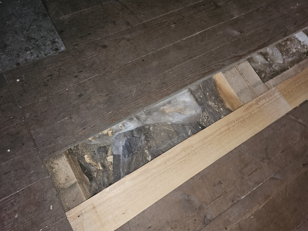 Offener Bodenspalt mit Holzbalken, Staub und Dämmmaterial sichtbar.