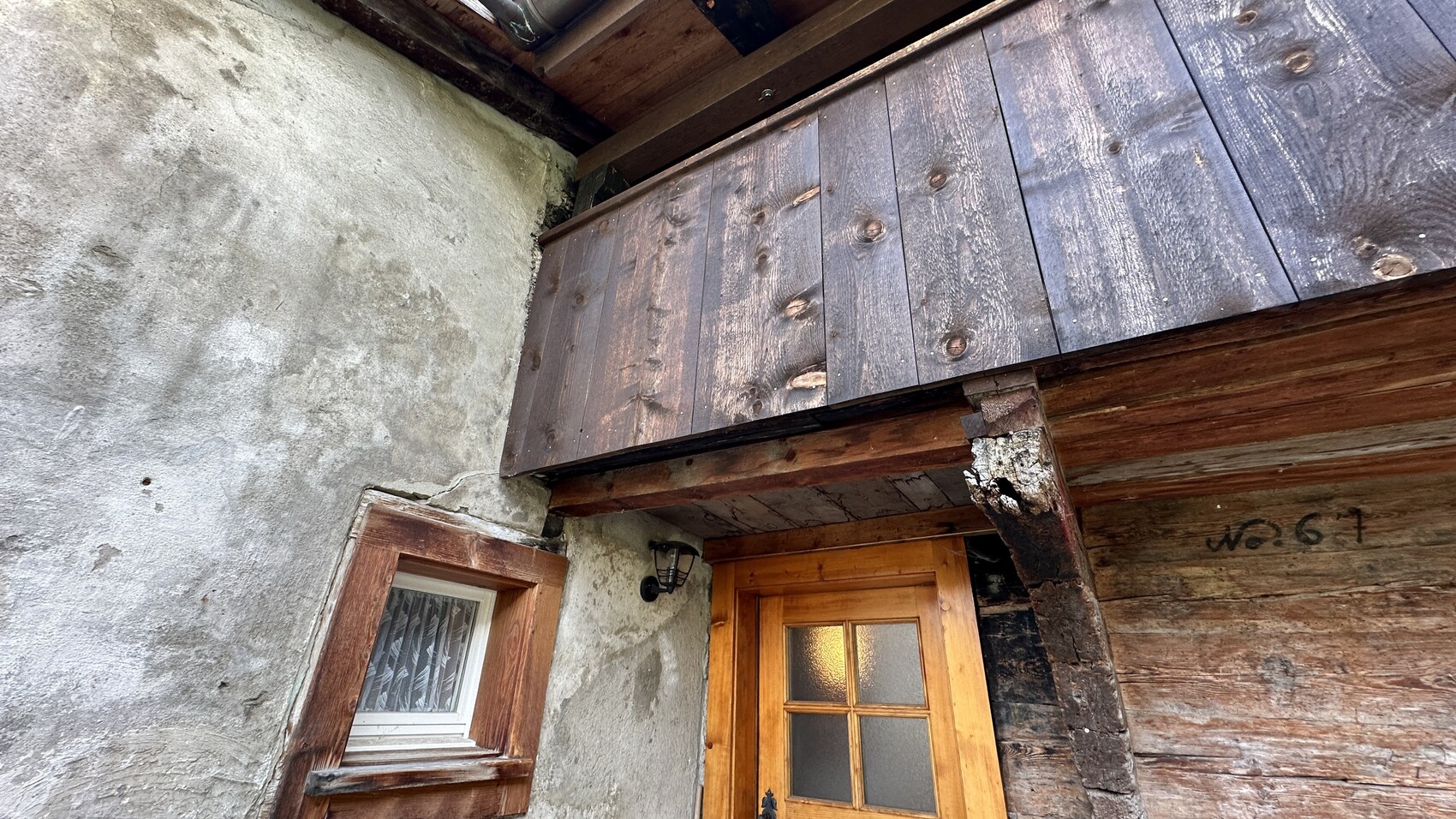 Außenansicht eines alten Holzhauses: dunkle Holzwand, Balkenüberbau über Tür.