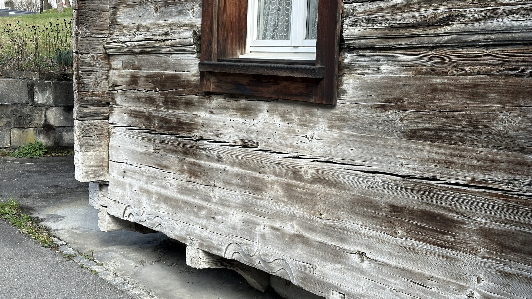 Verwitterte Holzwand eines alten Hauses mit kleinem Fenster und sichtbarem Fundament.