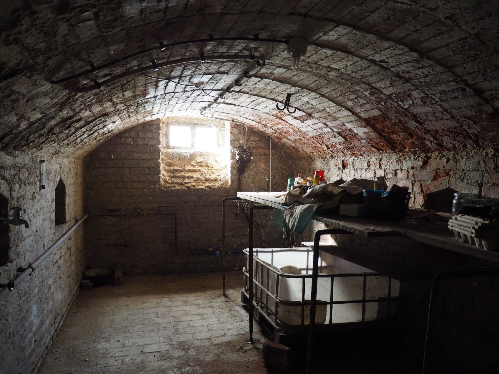 Keller mit gewölbter Backstein-Decke, kleinem Fenster und Tisch voller Utensilien und Behälter.