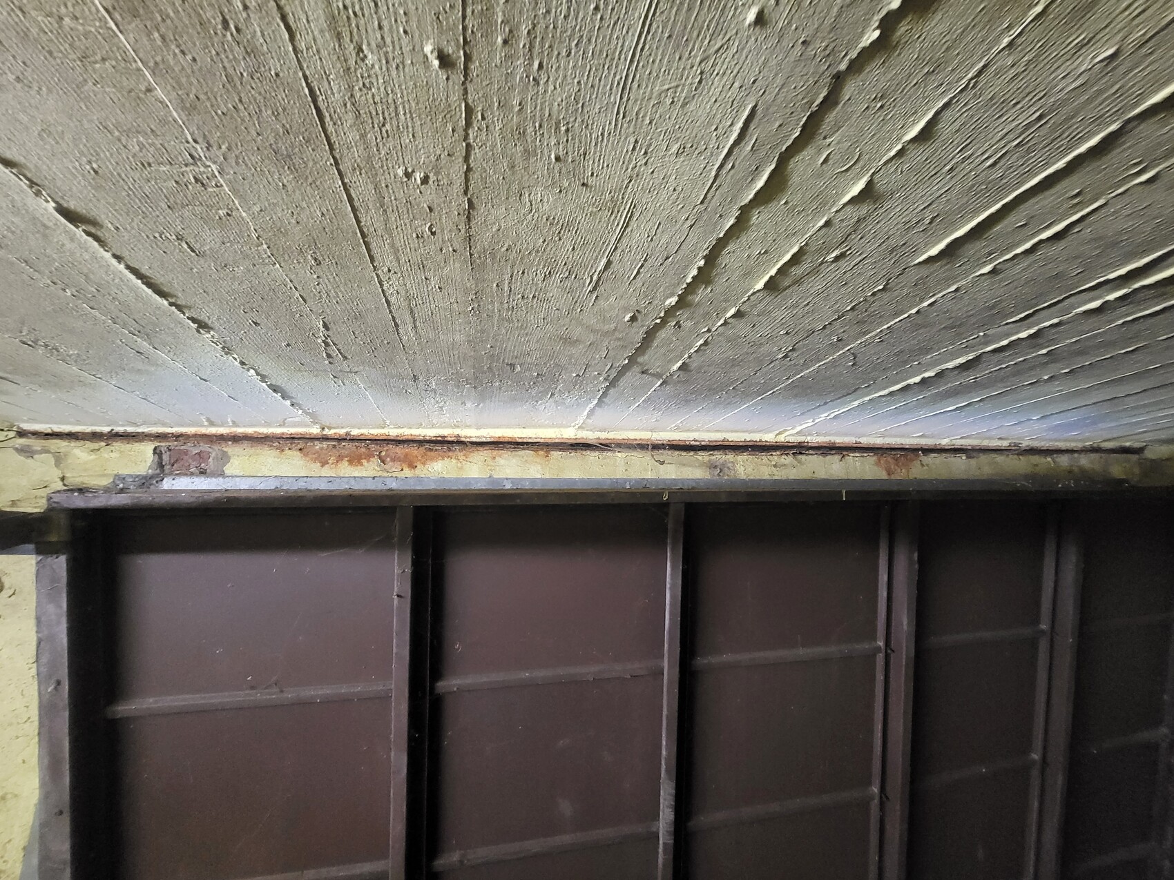 Untersicht einer grob gestrichenen Betondecke mit rostiger Metallleiste über braunen Wandpaneelen.