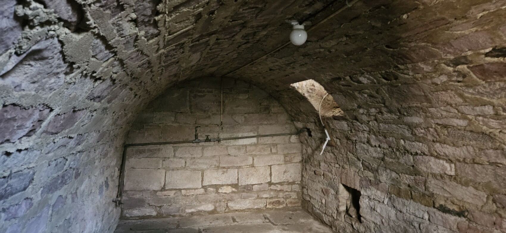 Dunkler Stein-Gewölbekeller mit Bogen, sichtbare Rohre, Glühbirne an der Decke, rechte Wandöffnung.