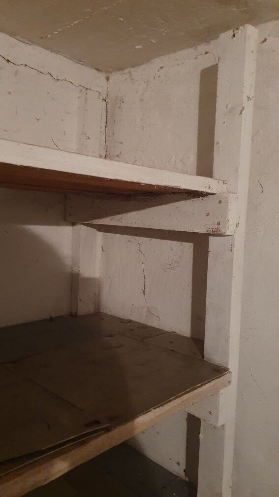 Verstaubte weiße Holzregale in einer kellerartigen Nische mit Rissen in der Wand.