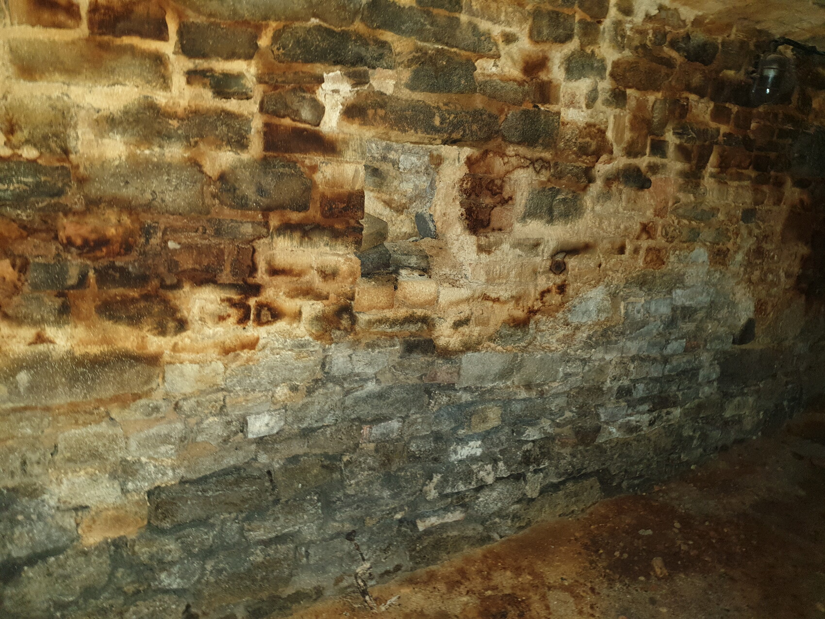Unverputzte Steinmauer im Keller: grobe Bruchsteine, feuchte Wand, erdiger Boden, warmes Licht rechts.