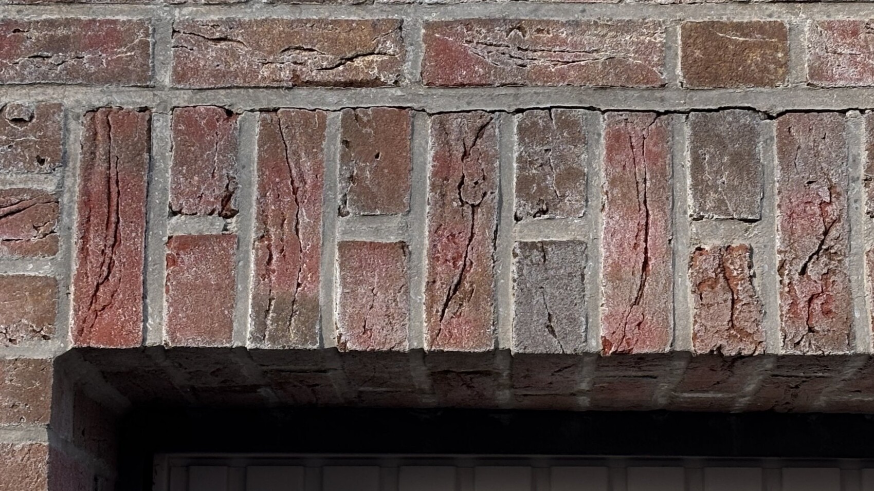 Rote Ziegelwand über einer dunklen Türöffnung, grobe Fugen und sichtbare Risse im Mauerwerk.