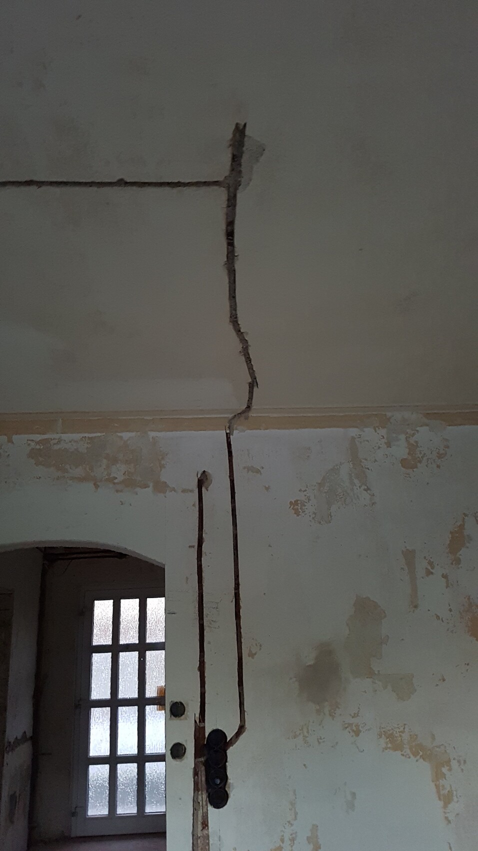 Verfallene Innenwand mit Deckenriss und rostigen Rohren; Tür mit Glasfenstern im Hintergrund.