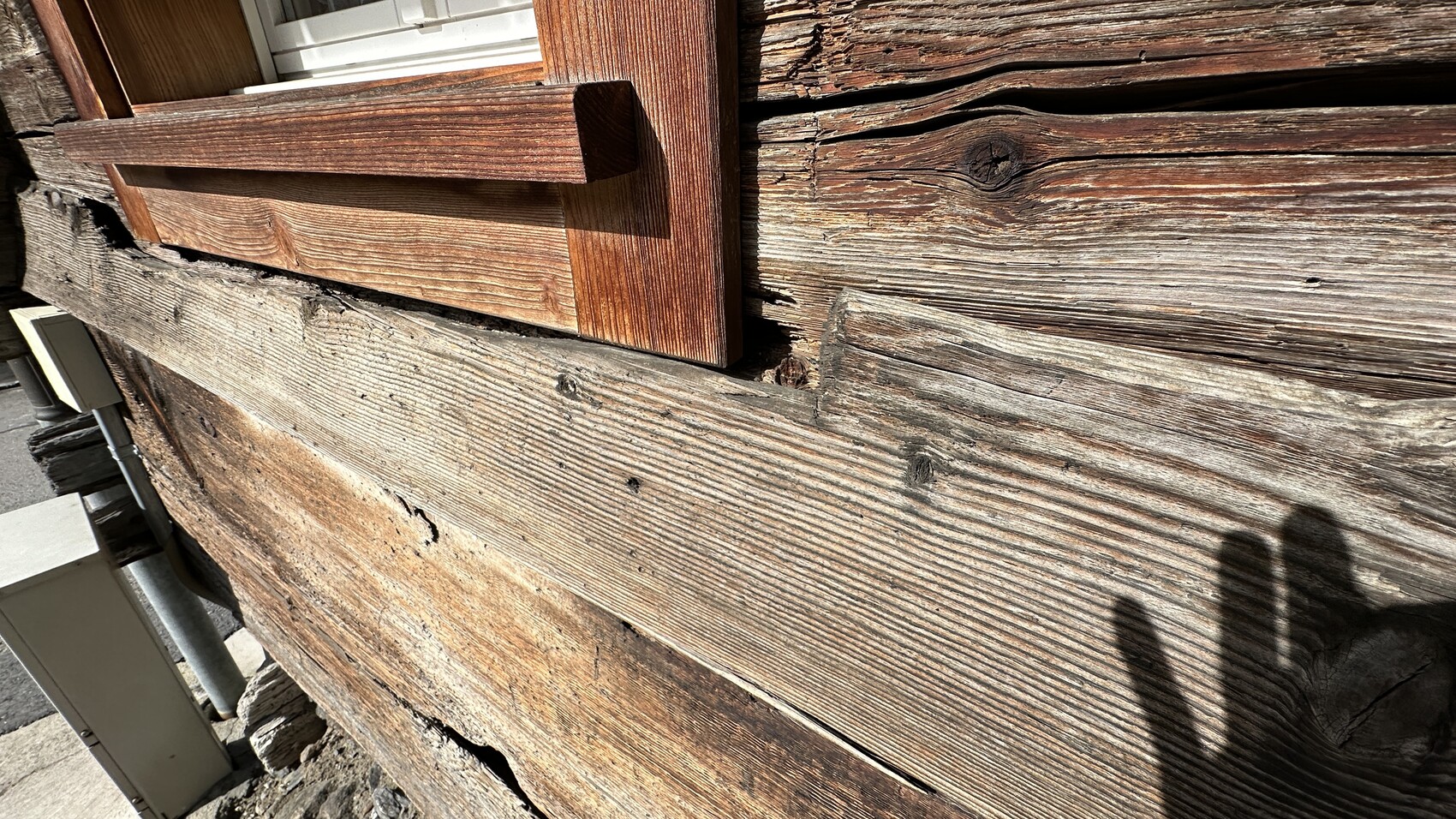 Nahaufnahme einer verwitterten Holzwand mit dicken Brettern, Fensterrahmen und Schatten einer Person.