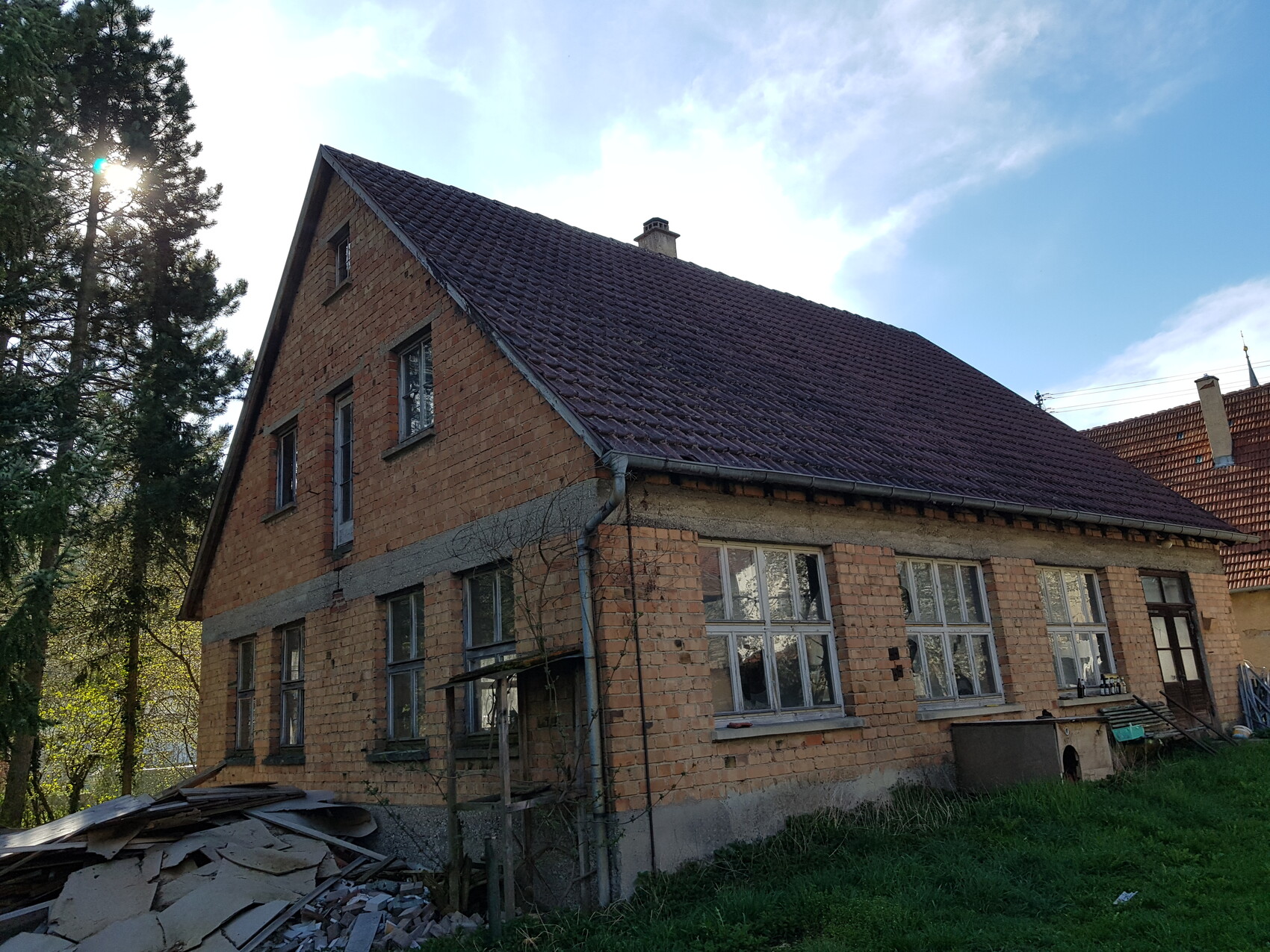 Altes Ziegelhaus mit Schraegdach, Bauabfall im Vordergrund und umliegenden Bäumen.
