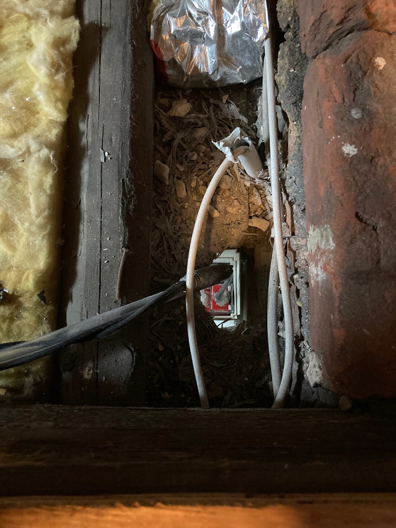 Freigelegte Kabel in staubiger Wandöffnung zwischen Ziegel und Holz; Dämmung links, rotes Innenfach sichtbar.