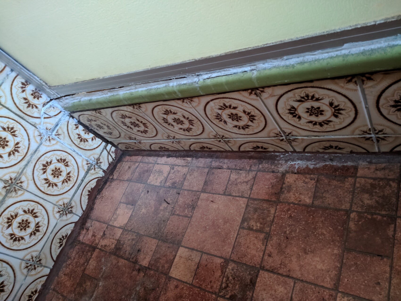 Ecke eines verdreckten Raums: grüne Wand, gemusterte Wandfliesen, lose Kabel, staubige Bodenfliesen.