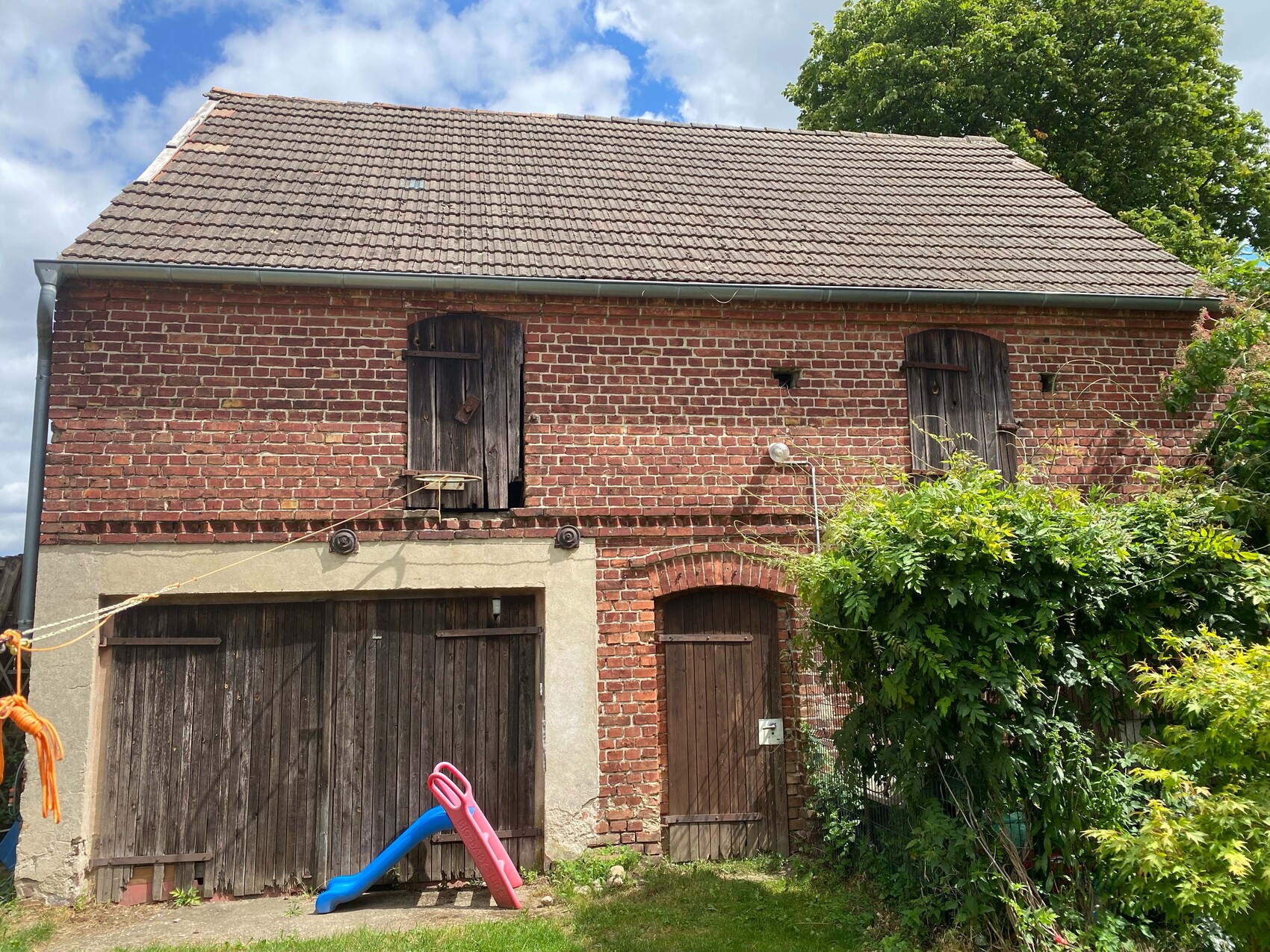 Altbau aus Ziegeln mit Holztüren, verschlossenen Fensterläden und bunter Spielrutsche im Garten.