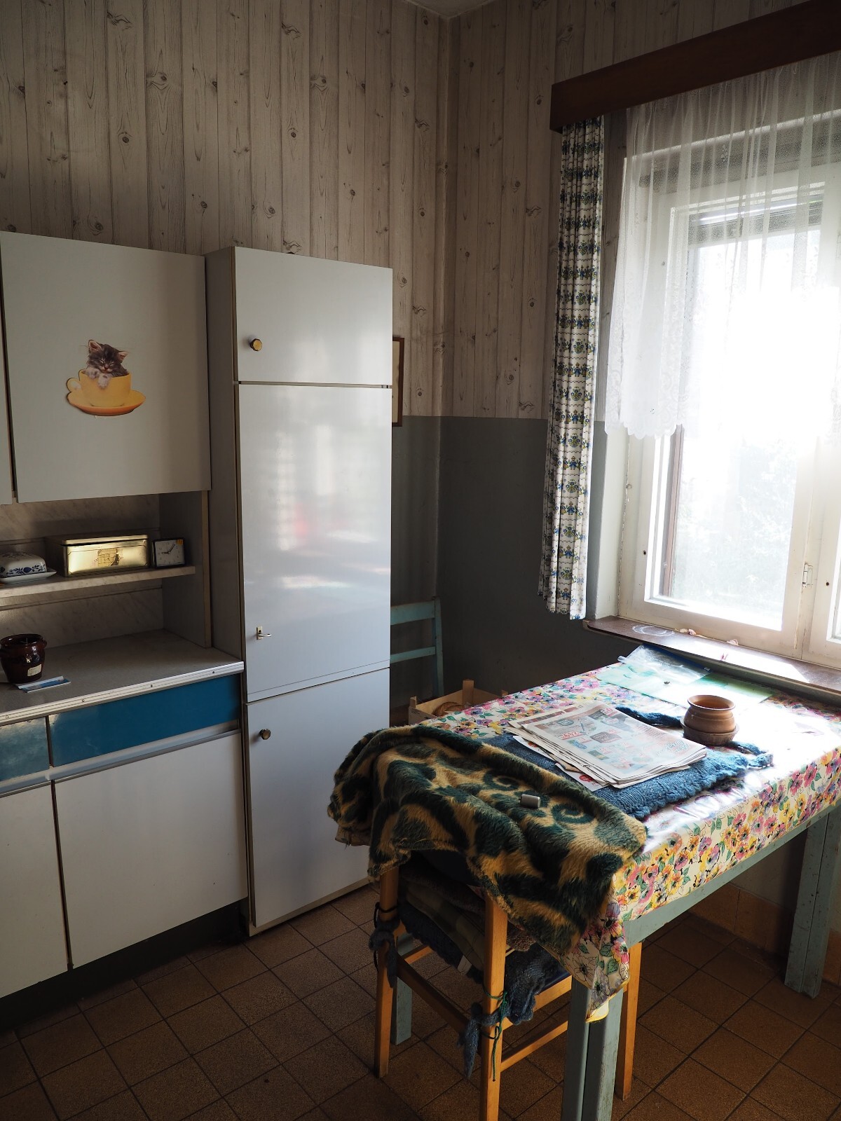 Küche mit weißem Kühlschrank, Katzenaufkleber am Schrank, Tisch mit Blumen-Tischdecke und Fenster.