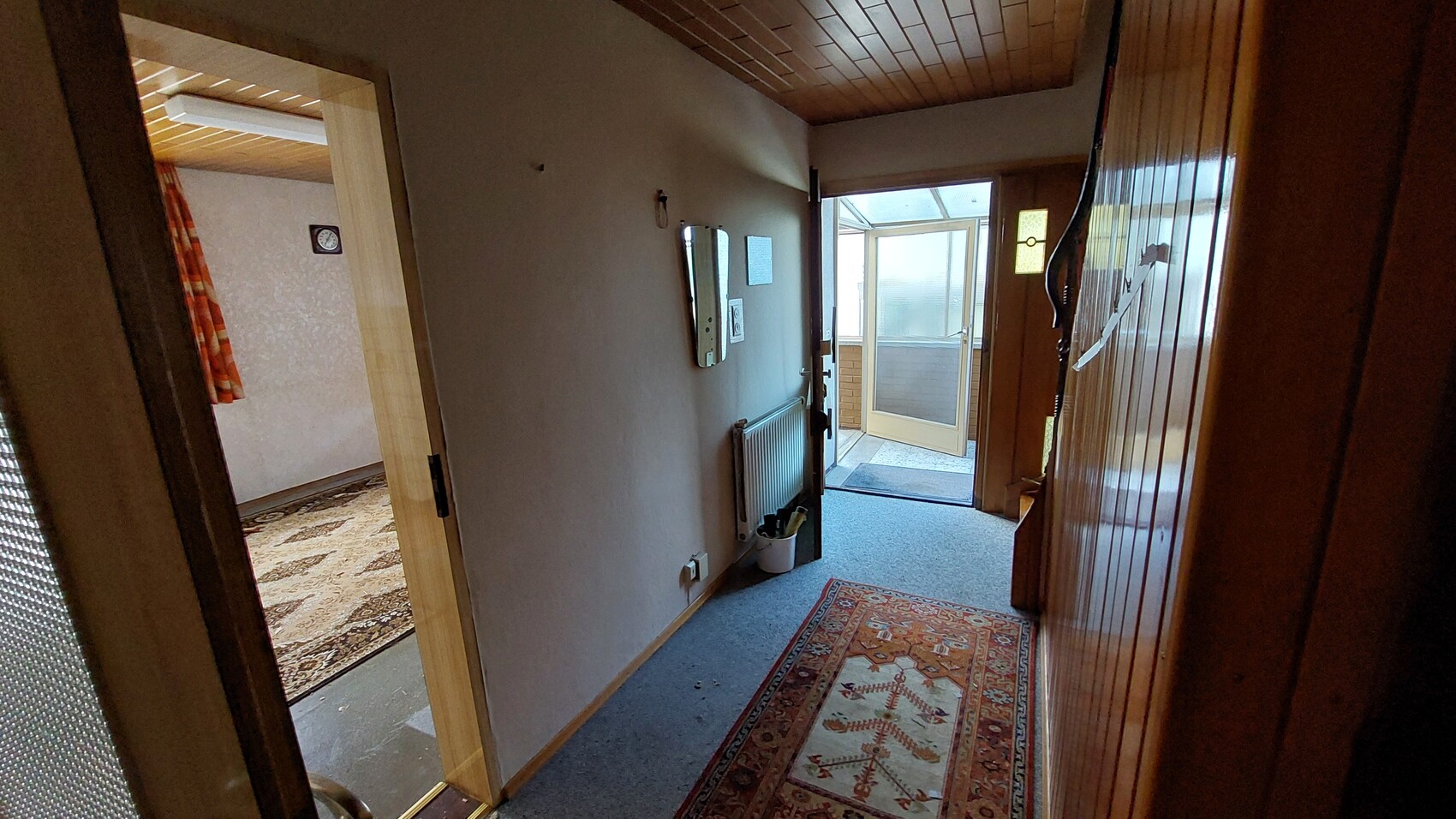Heller Flur mit Holzboden und Decke, linkes Zimmer mit Musterteppich und Vorhang, Glastür am Ende.