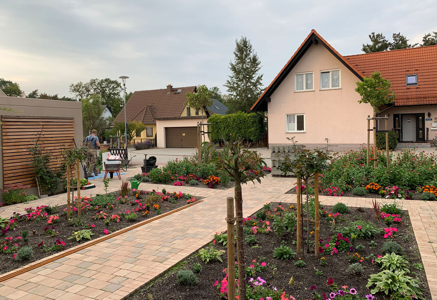 Gartenweg mit Blumenbeeten; links Person in Arbeitskleidung neben Schubkarre; Häuser im Hintergrund.