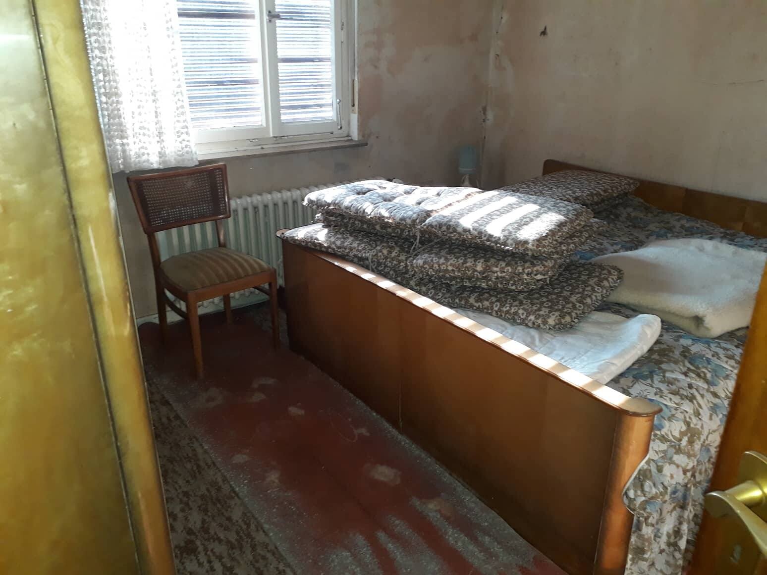 Altes Schlafzimmer: Holzbett mit gestapelten Decken, Stuhl, Fenster mit Vorhang und Radiator.