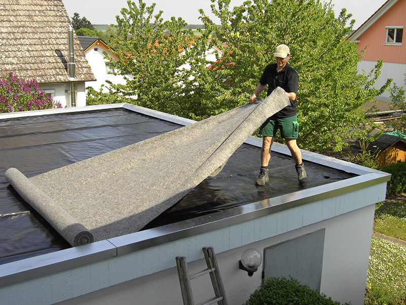 Mann rollt Dachpappe auf ein Flachdach eines Wohnhauses; Leiter steht daneben.