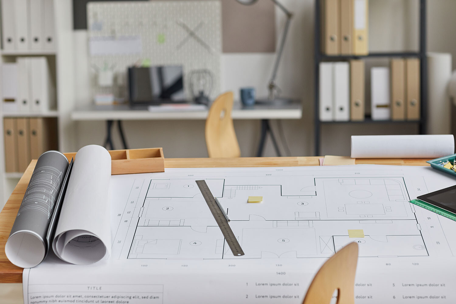 Architekturplan auf Schreibtisch mit Lineal und Rollpapier; Büro im Hintergrund