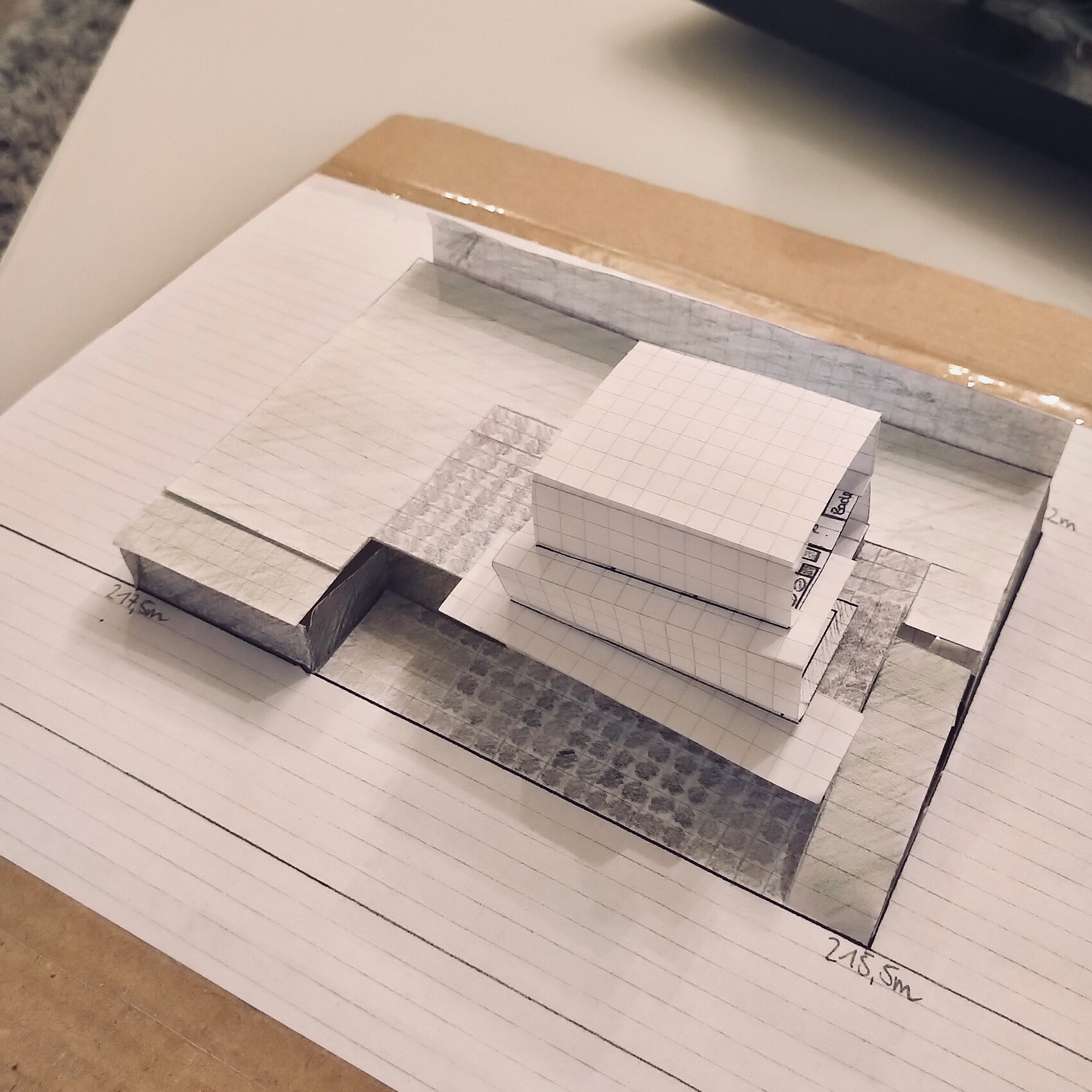 Papier-Architekturmodell mit gestapelten quadratischen Blöcken auf Gitterpapier-Grundriss.