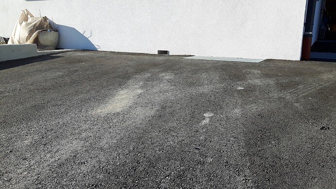 Asphaltierter Vorplatz vor weißer Hauswand, Eingang rechts, links Bauabfälle.