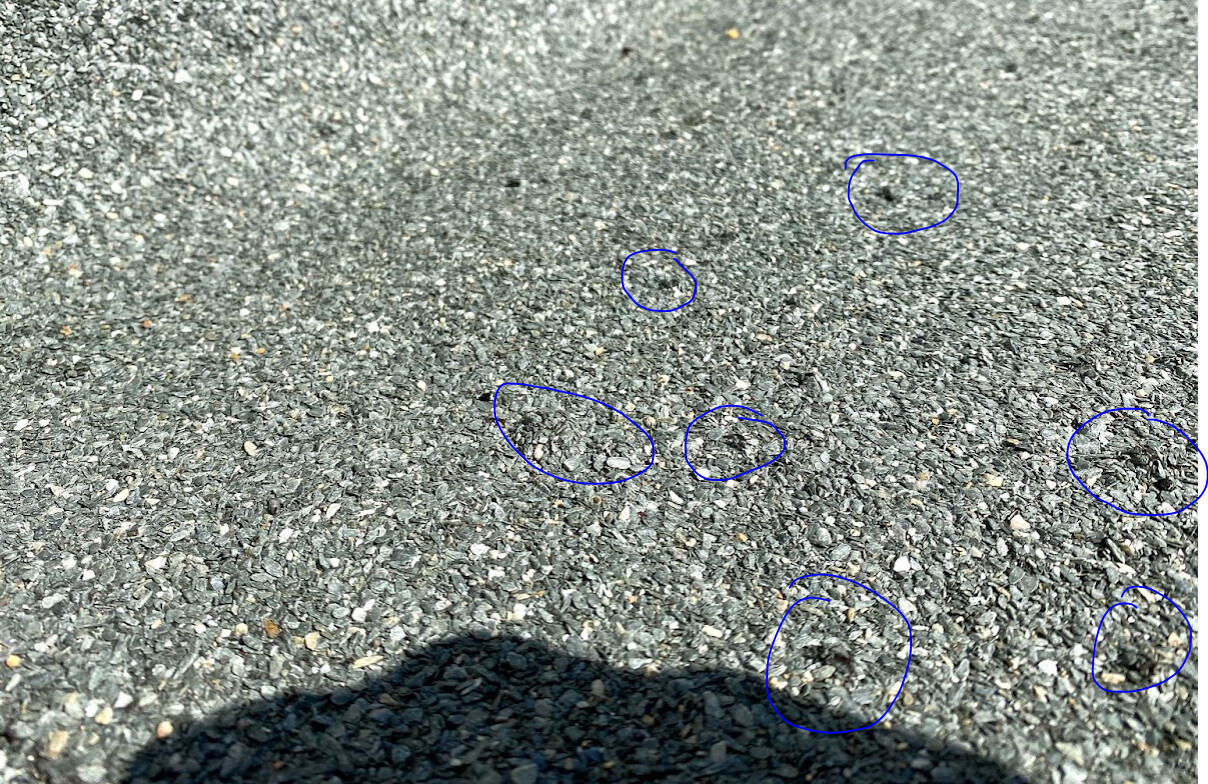 Asphaltfläche mit Kieselsteinen; blaue Kreise markieren Steine; Schatten einer Person unten links.