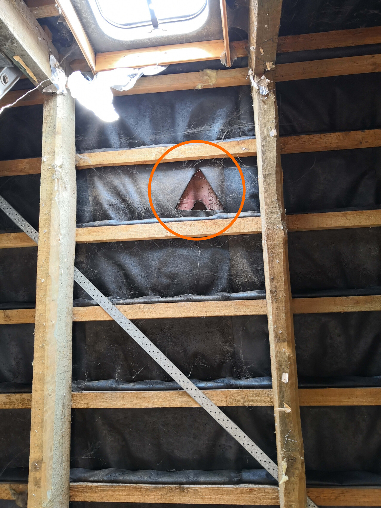 Dachboden mit Holzbalken und dunkler Dämmfolie; dreieckiges Loch zeigt roten Ziegel dahinter.